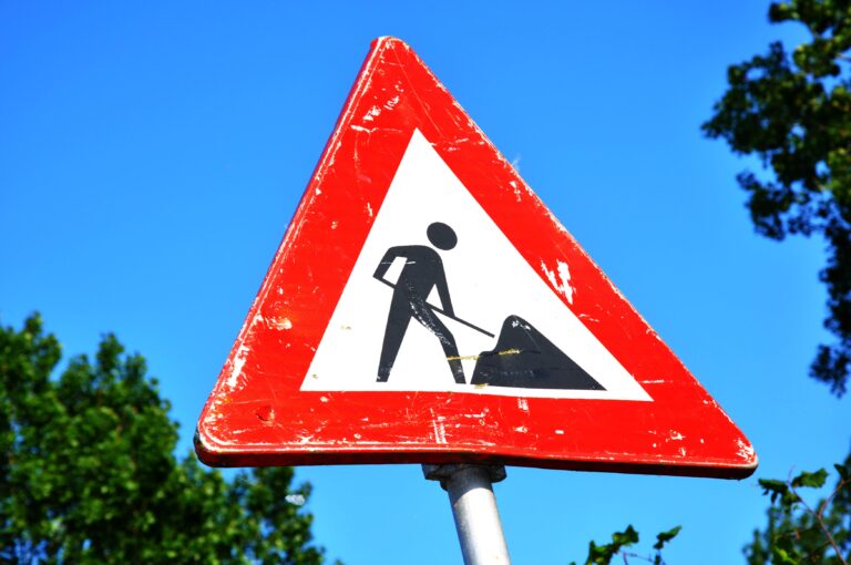A red and white roadwork warning sign with a silhouette figure, set against a clear blue sky.